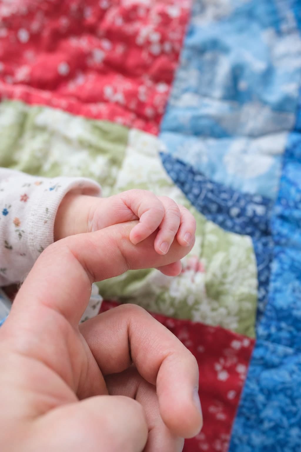 A newborn's hand curled around an adult's finger on a patchwork quilt